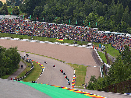 Motorrad Grand Prix Sachsenring 2025 Blick auf eine Kurve des Sachsenrings beim Motorrad Grand Prix Deutschland mit dicht besetzten Zuschauerhängen und mehreren Rennmaschinen im Pulk auf der Strecke – die große Fanmenge zeigt die besondere Atmosphäre der traditionsreichen Rennstrecke in Hohenstein-Ernstthal.